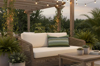 A rectangular decorative throw pillow with a textured, woven geometric pattern in shades of olive green, and blues, set against a plain white background. Finished in a Light Blue Trim. Outdoor setting on bench cushions.
