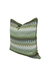 A square decorative throw pillow featuring a woven geometric chevron pattern in shades of olive green, sage, and blues, set against a plain white studio background.