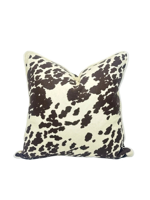A square throw pillow featuring a dark brown cow print pattern on a cream background, set against a plain white studio backdrop.