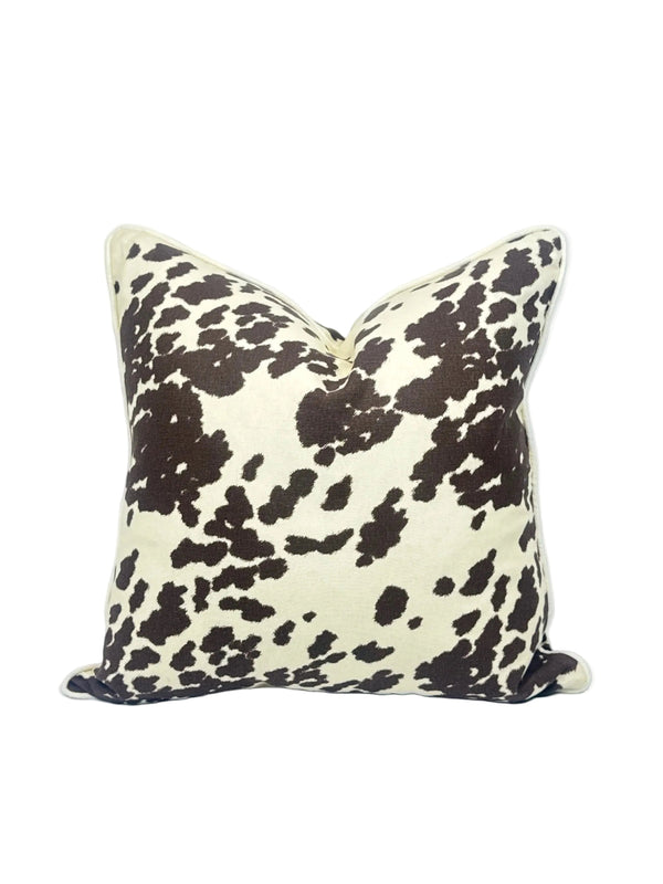 A square throw pillow featuring a dark brown cow print pattern on a cream background, set against a plain white studio backdrop.
