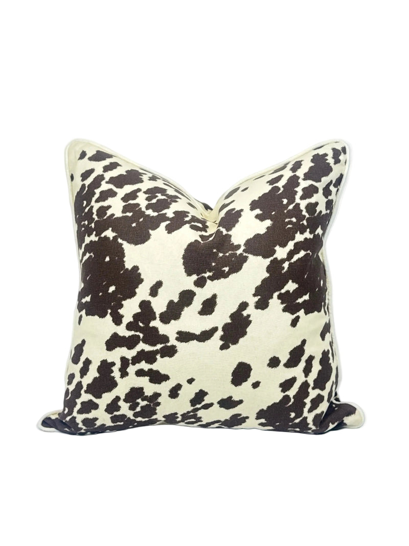 A square throw pillow featuring a dark brown cow print pattern on a cream background, set against a plain white studio backdrop.