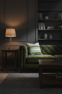A square decorative throw pillow featuring a vertical chevron pattern in shades of olive green, sage, and blues. The pillow is on a Green velvet couch in a moody room with built-ins against a dark grey wall. 