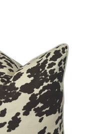 A close-up of a decorative throw pillow with a brown cow-print pattern on a cream background, shown against a plain white screen backdrop.